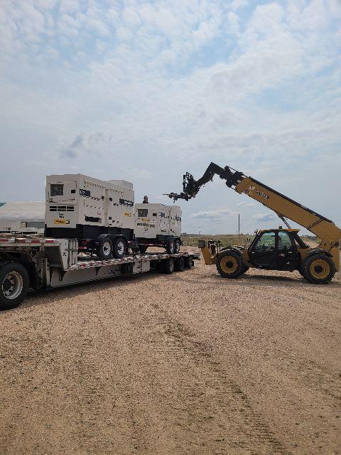 Two large portable generators loaded on a flatbed trailer with a yellow telescopic handler lifting one, in a desert-like area.