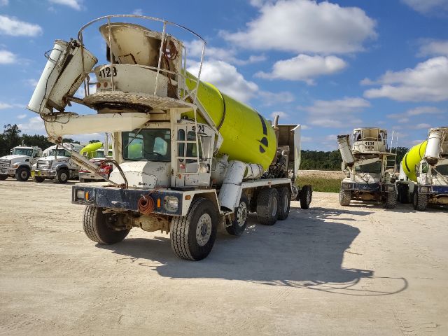 Concrete mixer truck with a yellow mixing drum in an outdoor yard; other mixer trucks in background under a blue sky.