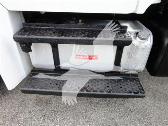 Step/footboard and fuel tank area of a Freightliner Business Class M2 106 showing a dual-step entry with a label on the tank and a perforated metal running board.