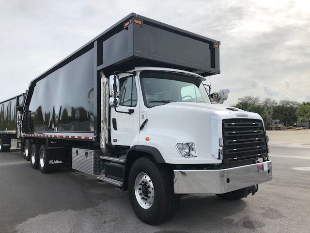 Front view of a white Freightliner 114SD grapple truck with a black dump body, showing large grille and dual rear axles. No visible text reads clearly other than generic truck features.