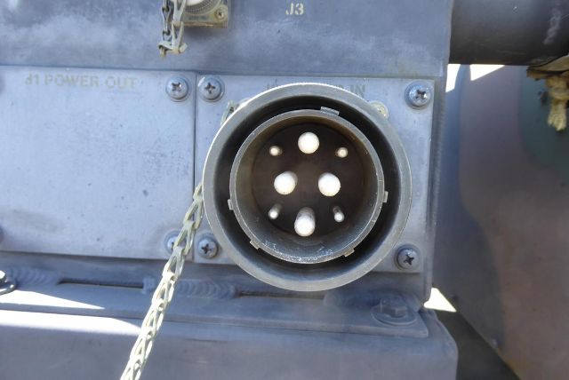 Trucking equipment power outlet showing a circular 5-pin electrical connector port with surrounding metal housing and chain nearby.