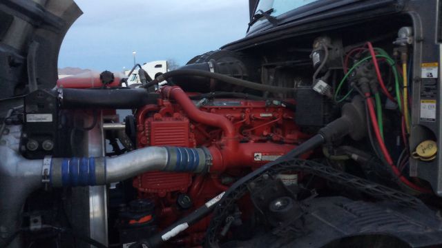 Engine bay of a Volvo truck showing a red engine block with blue-taped hose, silver intercooler pipe, various hoses, wiring harnesses, and battery/engine components.