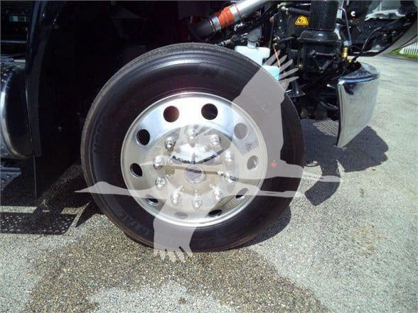 Front wheel and exposed chassis of a 2024 International MV tow truck, showing steel wheel with lug nuts and tire, and partial front bumper area.