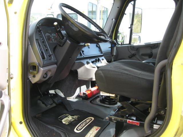 Image of a Freightliner M2106 cab interior showing steering wheel, dashboard, driver seat, pedals, and floor mats. Visible gear shift lever and inspection stickers near the door.