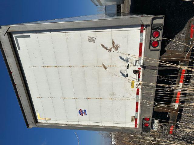Rear view of a white enclosed trailer with a roll door. Visible text includes ‘UTILITY’ logo sticker, small labels, and taillights. Grass and tall weeds in foreground.