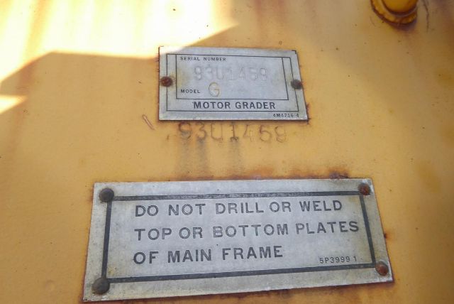 Plate labels on a yellow Caterpillar motor grader showing model and serial information. Upper plate reads SERIAL NUMBER 93U1458, MODEL G, MOTOR GRADER. Lower plate warns: DO NOT DRILL OR WELD TOP OR BOTTOM PLATES OF MAIN FRAME.