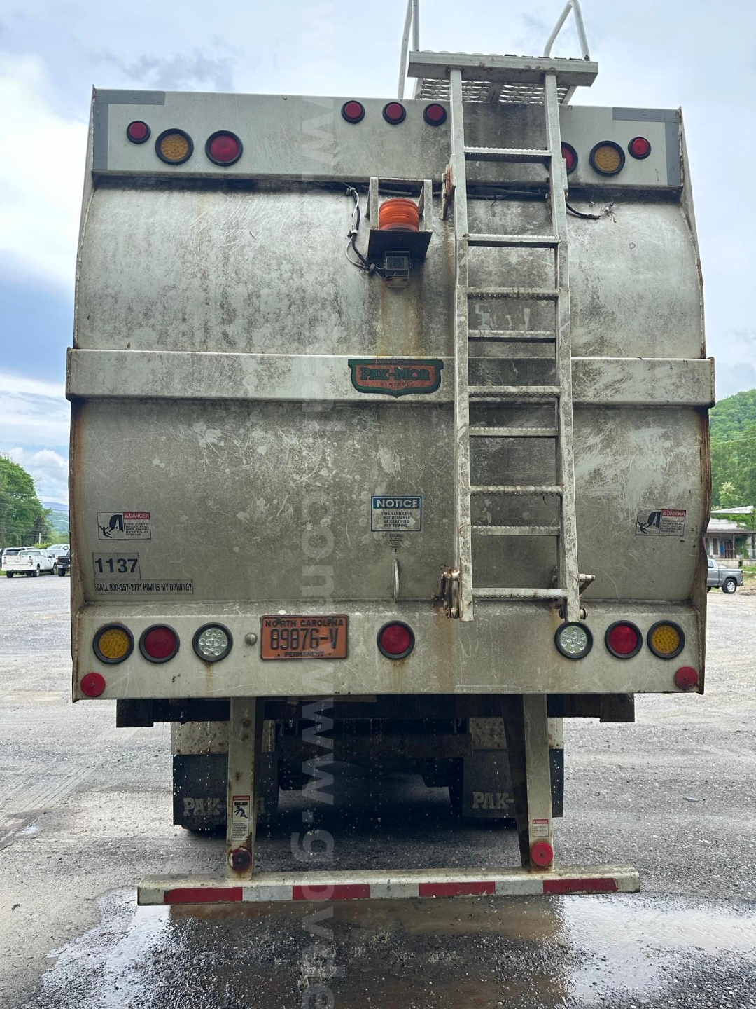 Back of a silver Mack MRU613 refuse truck with ladder, lights, and bumper stickers. Visible text includes license plate and various stickers.