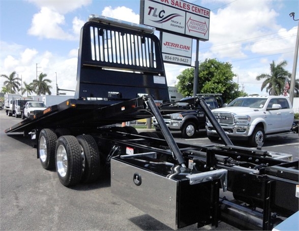 Kenworth W990 chassis with flatbed/tow equipment; visible labels include stock center signage in background, and truck showroom lot.