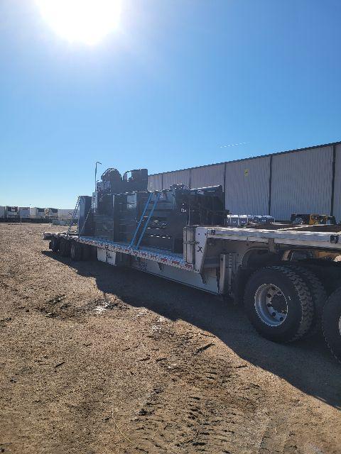 Flatbed trailer with equipment secured on it, visible tires and wheel well, and a large machinery load on the bed. Clear sky and building in background.