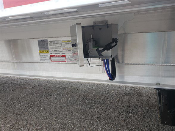 Trailer undercarriage showing hydraulic or electrical component box with label stickers and wiring harness along aluminum side rail.