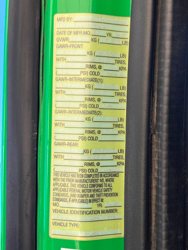 Tall yellow label with blanks for vehicle weights, GVWR, GAWR, rims, tires, and cold tire pressures on a green surface. Text includes MFD BY, DATE OF MFR, VEHICLE IDENTIFICATION NUMBER fields.