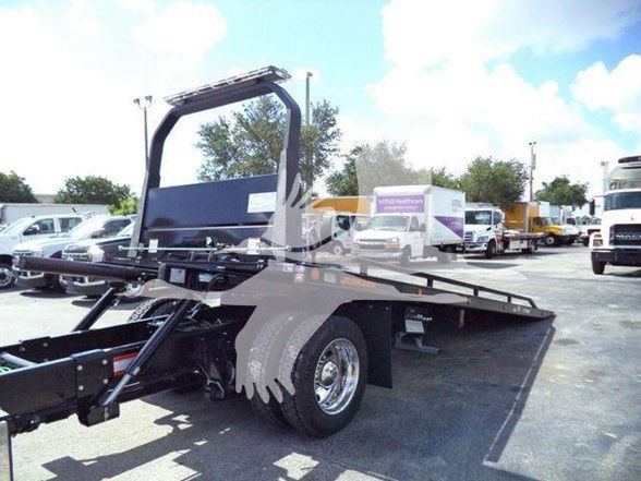 Tow truck chassis with flatbed and hydraulic tilting bed. Visible text includes a white truck with purple graphics in the background but no legible labels on the foreground equipment.