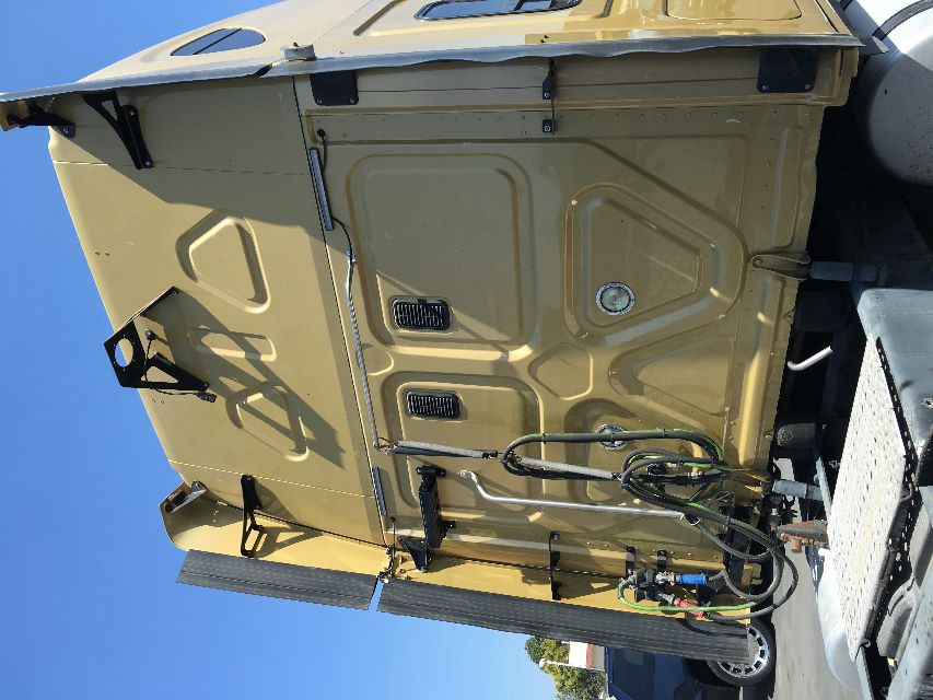 Rear view of a Freightliner Cascadia 125 sleeper tractor with beige cab, vertical exhaust, and various hoses hanging near the back door.