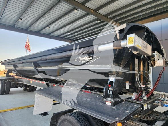 SmithCo dump trailer in-side view showing side-mounted hydraulic components, SmithCo logo, and black painted hopper on a low-boy trailer.