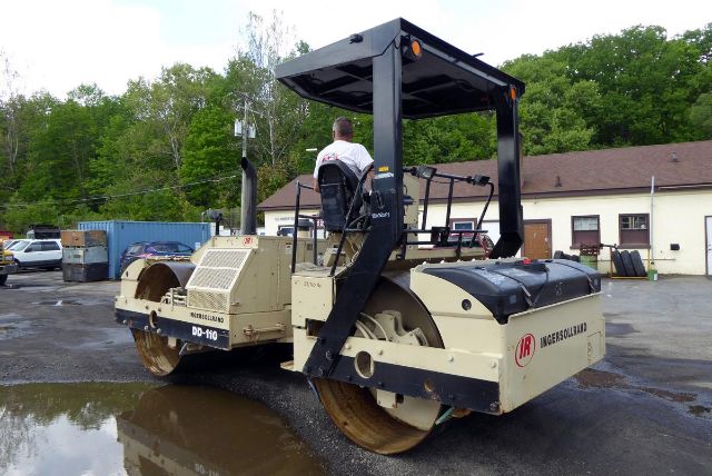 Ingersoll-Rand DD110 wheeled roller with operator seat and canopy, side view showing drum and beige/black body; yard with trees in background.