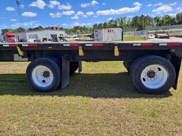 Flatbed trailer side view showing dual rear axles with white wheels, black frame, and reflective tape along top edge.