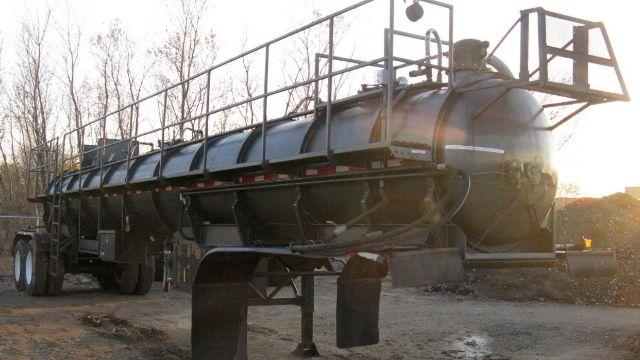 Vacuum tanker trailer in-service with large cylindrical tank, metal frame, rear platform, and landing legs. Tank appears black with walkway rails along top; dual rear axles with tires visible.
