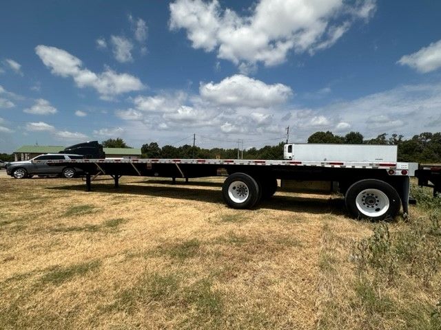 Utility flatbed trailer with dual axles and white rims, 48x102, in a field under a blue sky.