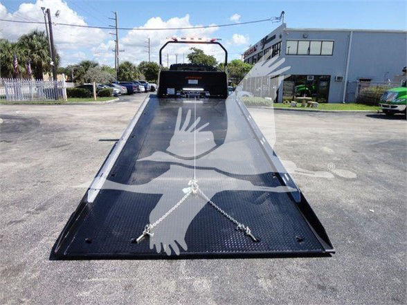 Tow truck flatbed with metal diamond-plate surface and safety chains, viewed from the front of the bed, with a handle and rear cab area visible.
