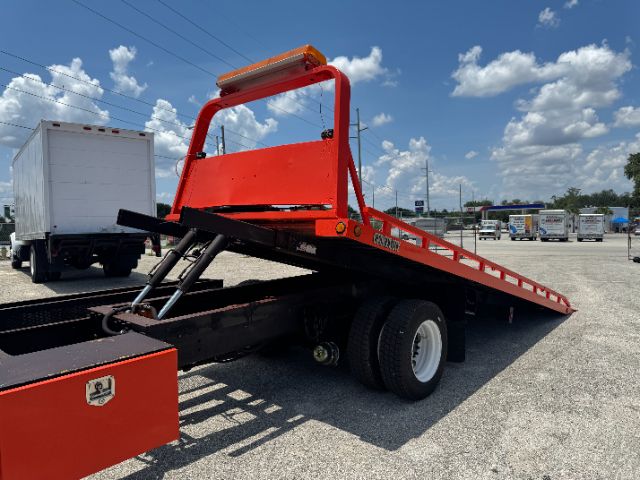 Utility heavy-duty tow truck flatbed with red equipment and hydraulic ramps; International 4700 DT 466E truck in background