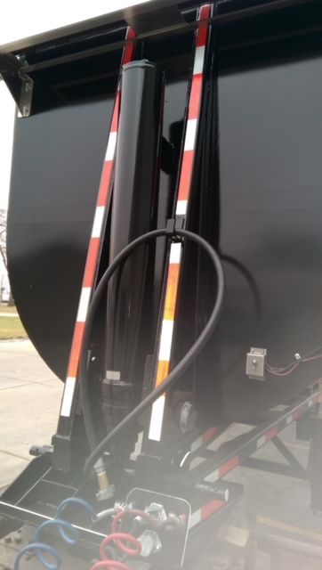 Dump trailer rear with round bottom; hydraulic cylinders and hoses visible, painted black with red/white reflective stripes.