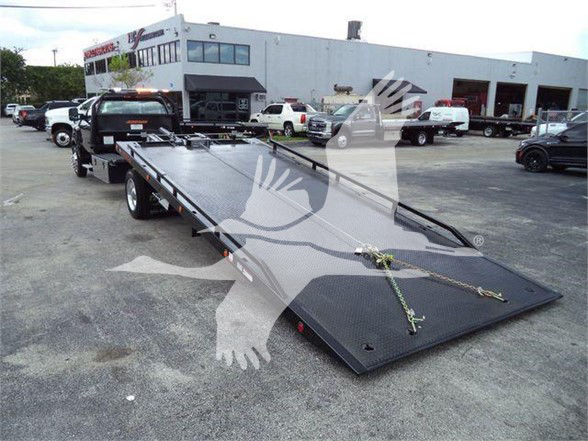 Tow truck with a long flatbed trailer in a dealership lot. Visible text includes stock number J56216 on a plate near the cab, and building signage in the background.