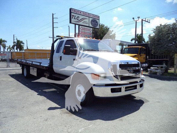 Flatbed tow truck (2015 Ford F-650) in a dealership lot, front three-quarter view showing white cab, long bed, and tow equipment.