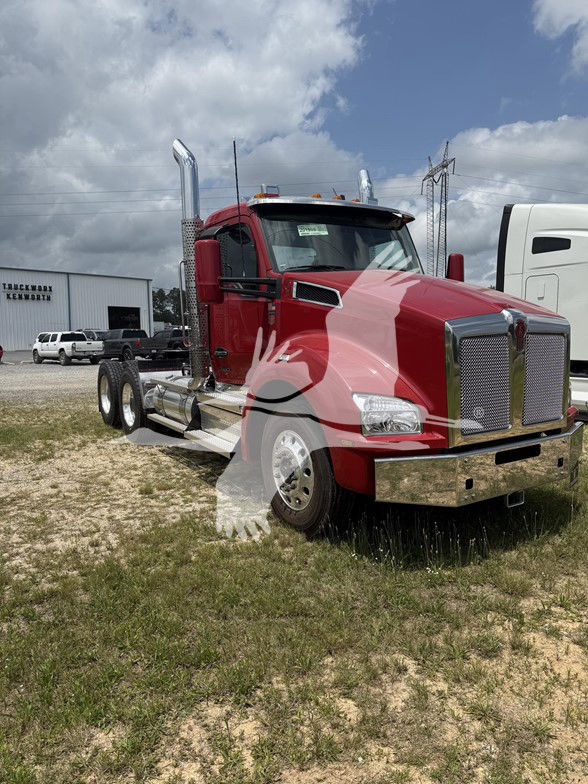 Red Kenworth T880 conventional daycab truck with chrome grille and exhaust stacks. Empty lot, other trucks in background.