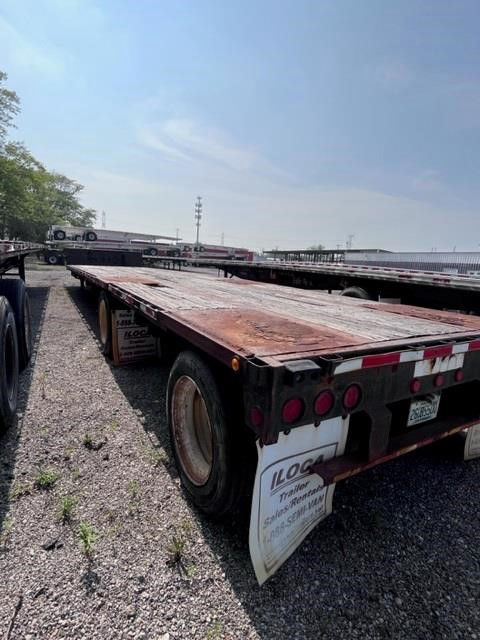 Flatbed trailer with weathered wooden deck, rear lights, and mudflaps. Some distant trailers visible in lot. No legible text prominent other than small sticker shapes.