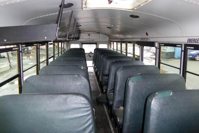 Bus interior with rows of dark green seats, narrow aisle, emergency exit sign visible along the cabin ceiling.