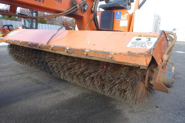 Trucking equipment with a large orange sweeper brush attachment. Visible labels on the top panel with warning text and mechanical linkage at right side.