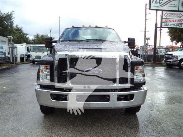 Front view of a 2024 Ford F-750 SD tow truck with large chrome grille, bumper, and Ford emblem; light bar on roof; dealership lot in background.