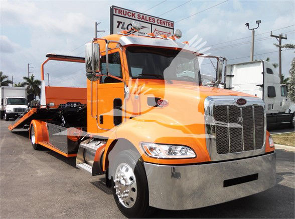 Orange Peterbilt tow truck in a dealership lot with a low chassis and chrome front bumper. Visible signs include 'Truck Sales Center' on a building in the background.