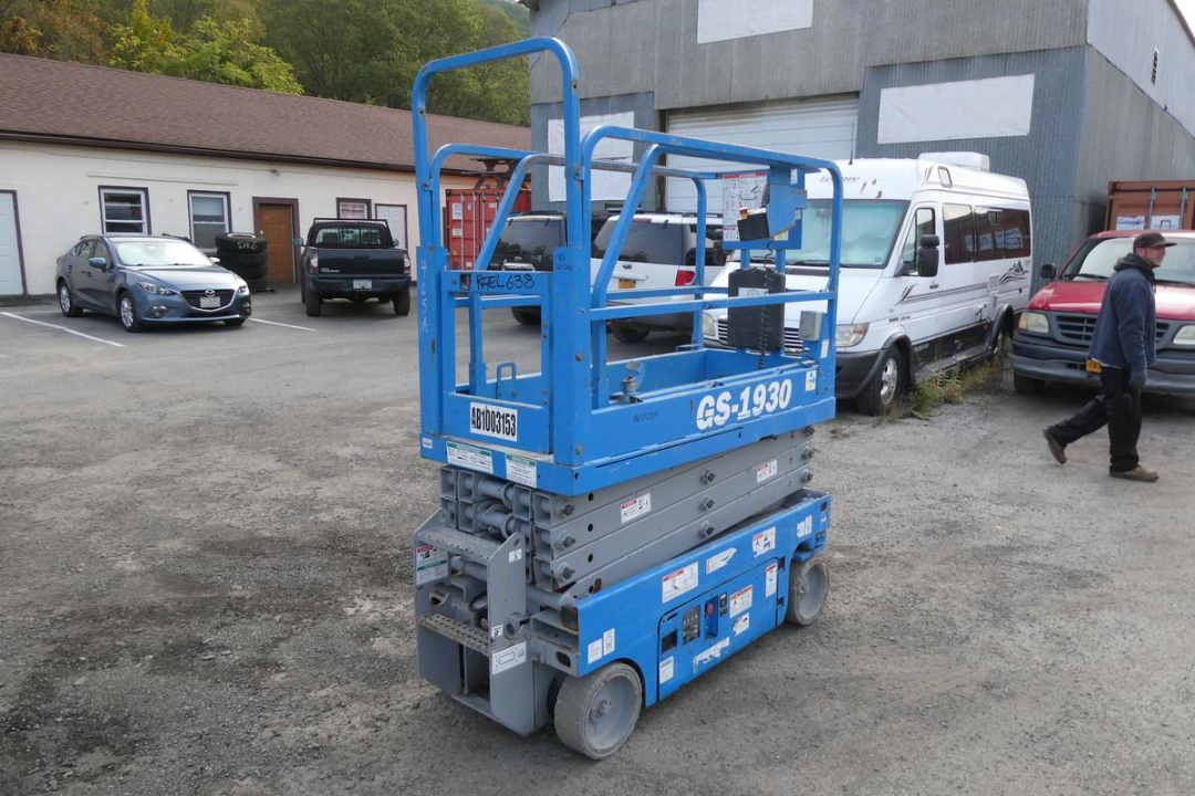 Scissor lift showing GS-1930 model branding, blue compact electric aerial work platform with guard rails and control panel. Side view reveals model text GS-1930.