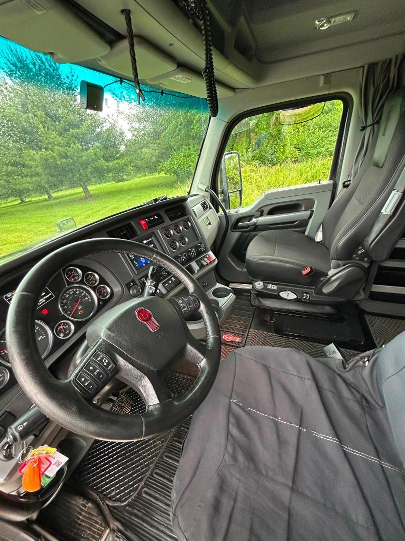 Kenworth T680 truck interior showing steering wheel, dashboard gauges, and driver seat; keys and accessories on lower left, gray seat cover, and windshield with green outdoor scenery.