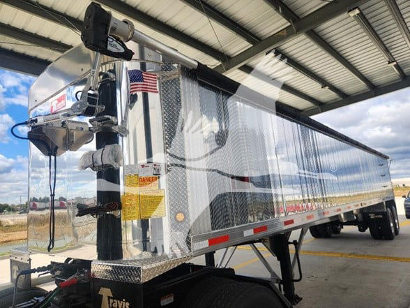 Travis dump trailer with shiny reflective diamond plate sides. Front-mounted hitch and electric crank system, warning labels on the front, American flag sticker, underbelly and rear wheels visible.