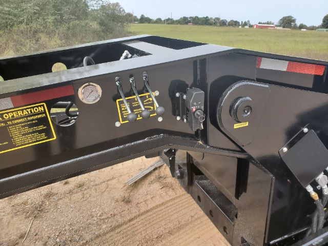 Trailer hitch area showing control levers, hydraulic reservoir gauge, and warning labels on a lowboy trailer.