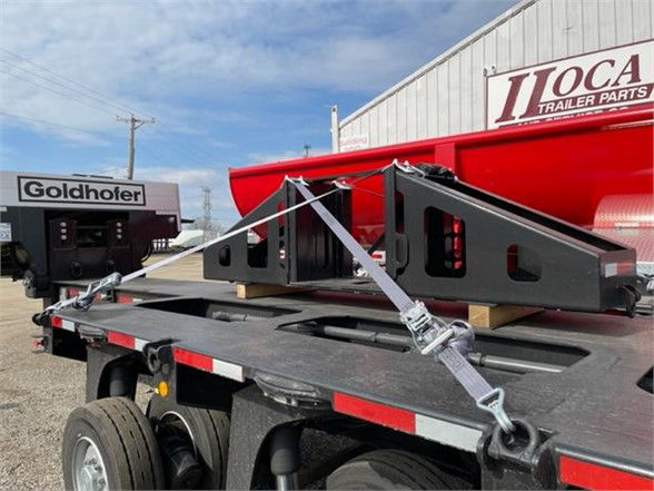 Flatbed trailer with a black steel triangulated rack and load straps on the bed. In the background is a red building with 'J&K Trailer Parts' sign and a white 'Goldhofer' sign on another trailer.