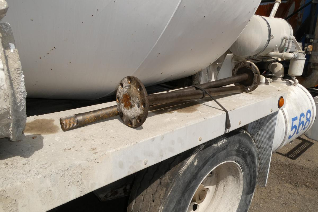 Axle area of a mixer truck showing a rusted axle shaft with flanged coupling and hoses on a white tanker behind the rear wheel.