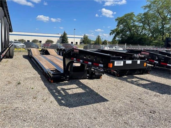 2023 Fontaine 55 TON steel roller paver lowboy trailer with 24ft deck. View shows flatbed sections, black frame, and some wheel wells with spare tires in background.