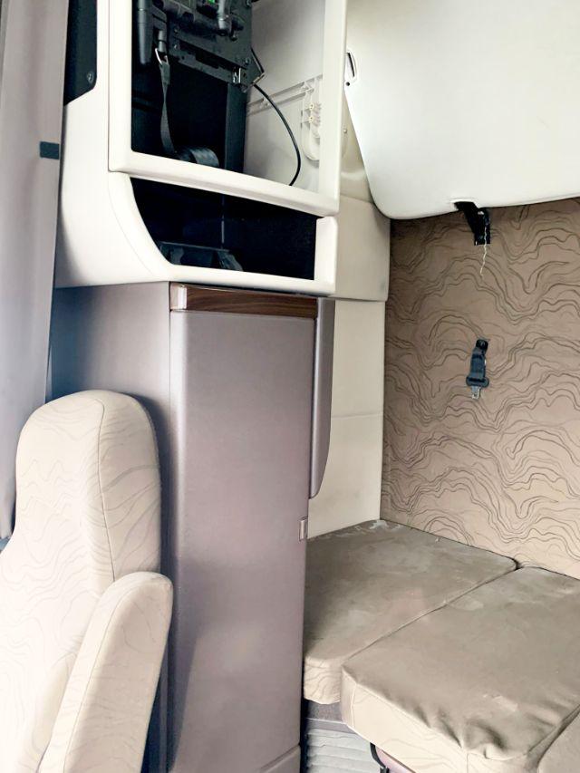 Truck cab interior showing storage area and sleeper bunk in a Volvo VNL 670 with beige upholstery and patterned wall panel.