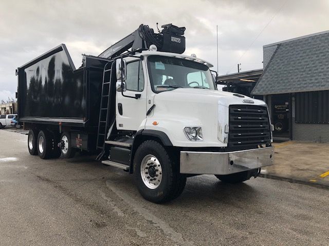 Truck in a grappler configuration: white Freightliner cab with large black grille, dump bed, and a crane on top.