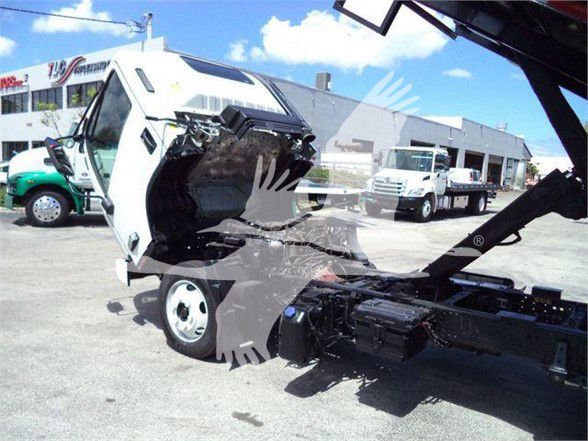 Isuzu NRR dump truck with cab tilted for engine access; front end raised, flatbed/arm lift visible in a dealership lot.