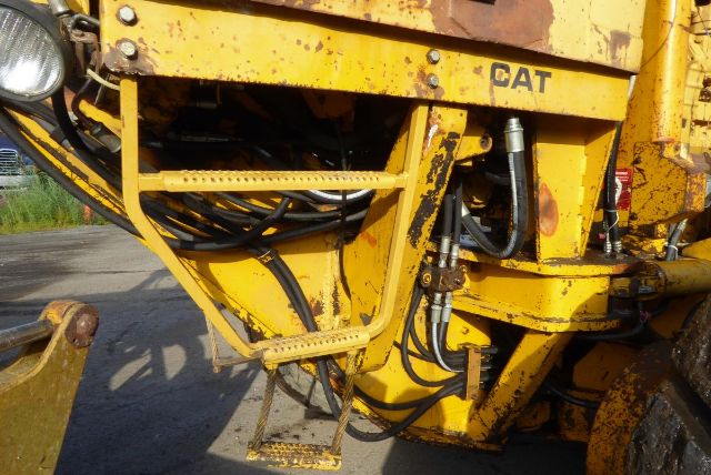 Caterpillar 16G motor grader component close-up showing hydraulic hoses, metal framework, and CAT logo on yellow-painted equipment.