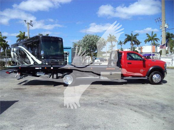 New red Ram 5500 Tow truck with a black car hauler trailer attached, large chrome wheel covers, palm trees and blue sky in background.