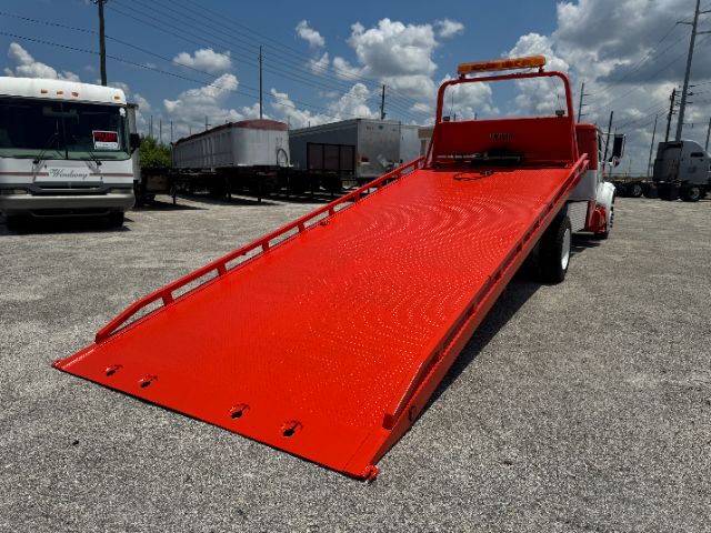 1999 International 4700 tow truck with bright red flatbed lowered for loading, DT 466E badge not visible, parked on gravel with other trailers in background.