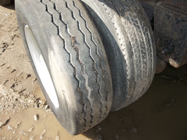 Dual rear wheels of a Wilson flatbed trailer showing treaded tires with worn patterns and mud; partial wheel rims visible.