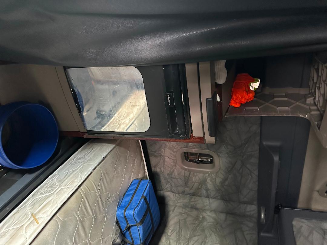Cab interior of a 2011 Freightliner Cascadia truck sleeper with bunk and storage, showing a small freezer/refrigerator and a blue bag.