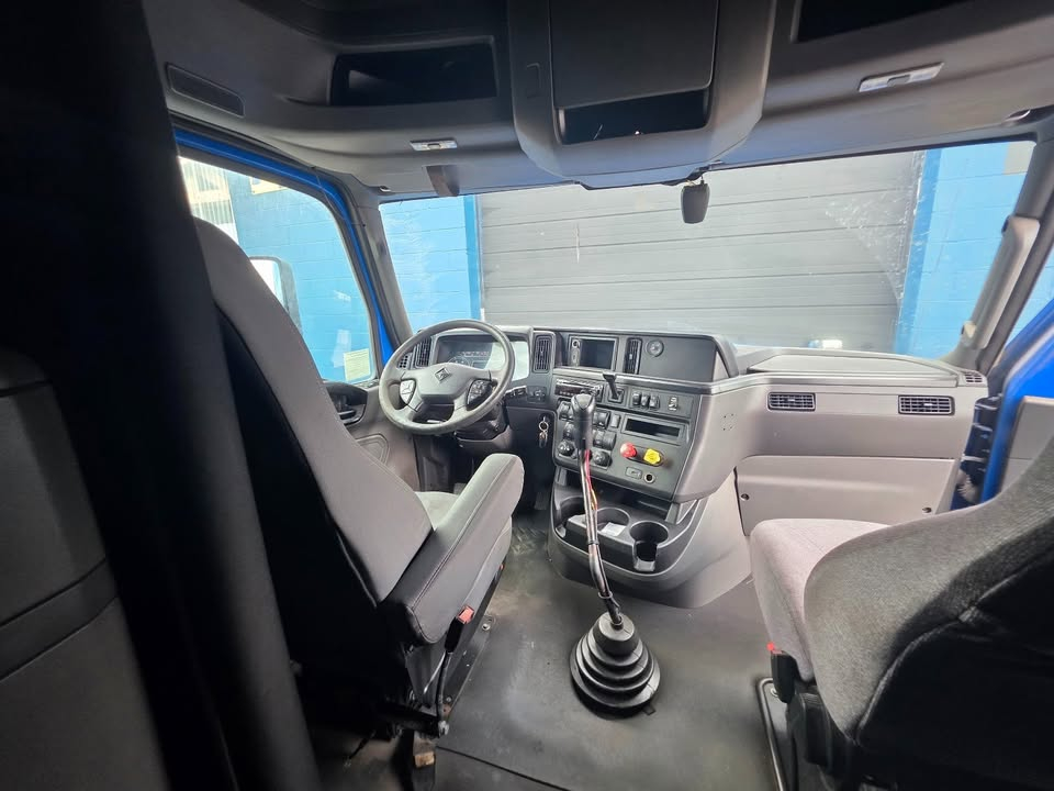 Truck cab interior showing steering wheel, dashboard, gear shift, and seating. Center console with controls and digital displays. Crew-style cabin with gray upholstery.