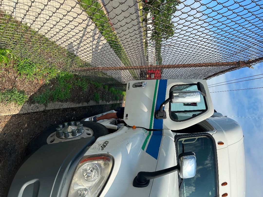 Truck cab of a 2011 Freightliner Cascadia with blue and green stripes, front left view showing headlight, large mirror, and wheel; chain-link fence in background.